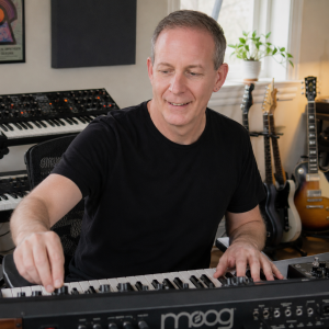 Robert Portillo adjusting a synthesizer in a home studio surrounded by keyboards and guitars